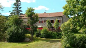 Un bâtiment en pierre, surmonté d’un toit de tuiles rouges, entouré d’une végétation luxuriante et d’arbres.