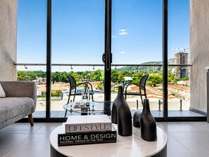 A modern living room with a large window, a sofa, a glass table with decorative items, and books on design trends. - Modern Studio in Art Haus San Benito in the City's Most Exclusive Area (San Salvador)