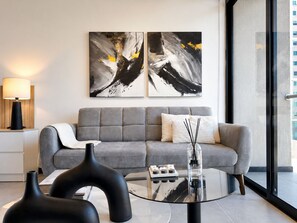 Ein modernes Wohnzimmer mit grauem Sofa, abstrakter Kunst an der Wand, einem Glastisch und einer Lampe auf einem Beistelltisch.