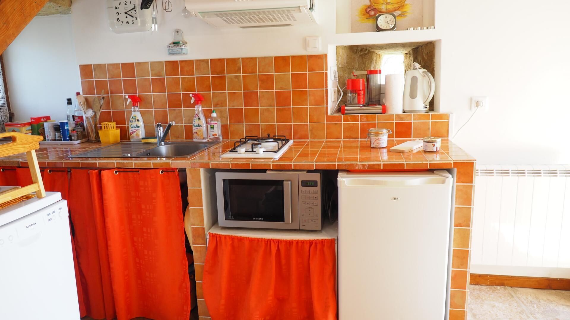 A kitchen with a microwave, a sink, and a small refrigerator. There are orange curtains, a clock, and a shelf with kitchen items.