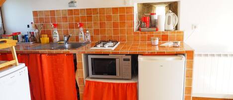 A kitchen with a microwave, a sink, and a small refrigerator. There are orange curtains, a clock, and a shelf with kitchen items.