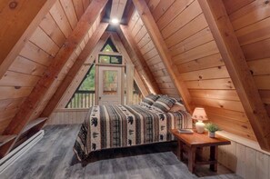 Ein gemütliches Schlafzimmer im Dachgeschoss mit schrägen Holzwänden, einem Bett mit gemusterten Textilien, einem kleinen Nachttisch mit Lampe und einem Fenster mit Blick auf Grünflächen.