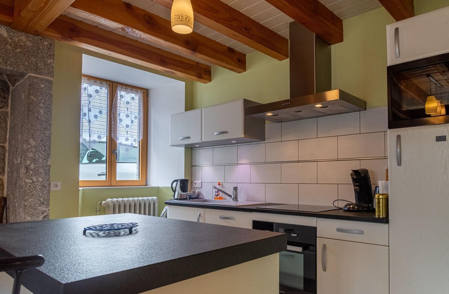Una cucina con soffitto in legno, mobili bianchi, piano di lavoro nero e finestra con tende a motivi.
