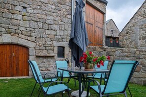 A stone building with a wooden door, a table with chairs, and a potted plant with red flowers. - Gîte de Montchamp - ARZENC D'APCHER (ARZENC D'APCHER)
