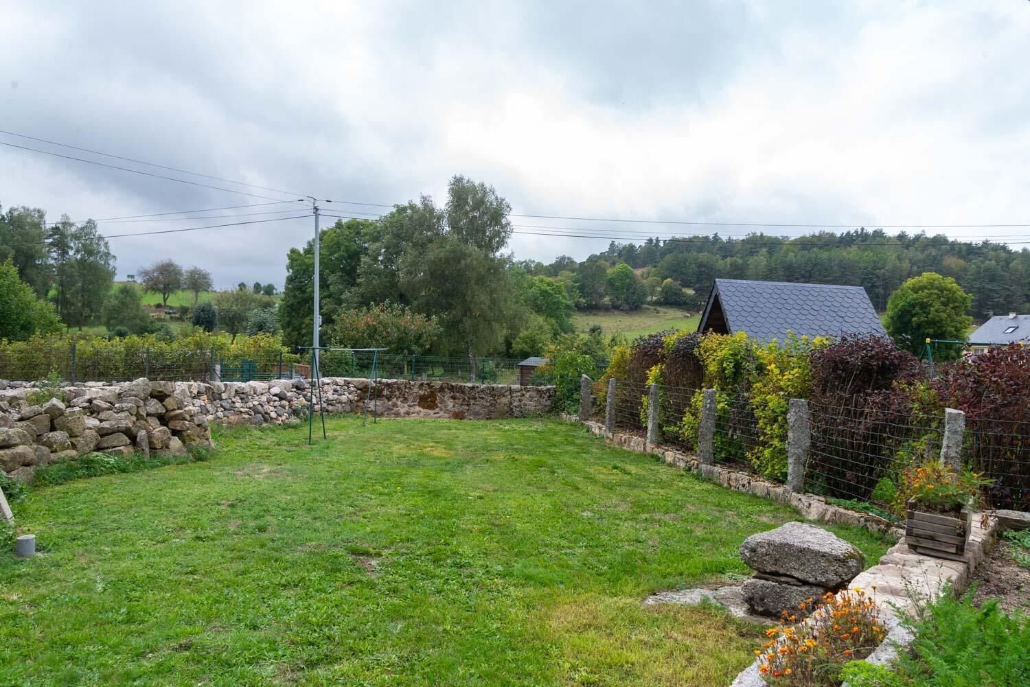 A rural backyard with a stone wall, a grassy lawn, and a house with a dark roof.