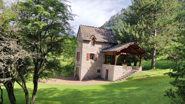 Una casa de piedra con porche de madera, rodeada de abundante vegetación y árboles.