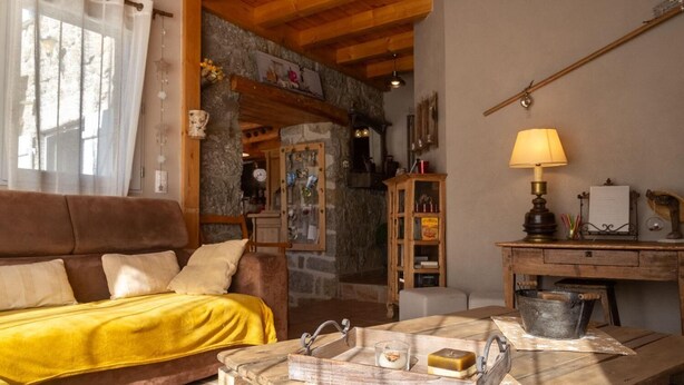 A cozy living room with a wooden ceiling, a brown sofa, a yellow throw, a wooden table with a lamp, and a stone wall. - L'Echappée de la Lozère - LES LAUBIES (LES LAUBIES)