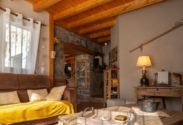 A cozy living room with a wooden ceiling, a brown sofa, a yellow throw, a wooden table with a lamp, and a stone wall. - L'Echappée de la Lozère - LES LAUBIES (LES LAUBIES)