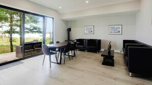 A modern living room with a dining area, black furniture, and large windows overlooking a garden. - Deluxe 3-Bedroom Cottage #12 (Nobel)