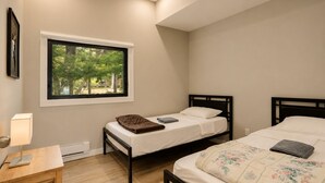 A bedroom with two beds, a window with a view of trees, a bedside table with a lamp, and a wooden floor. - Deluxe 3-Bedroom Cottage #12 (Nobel)