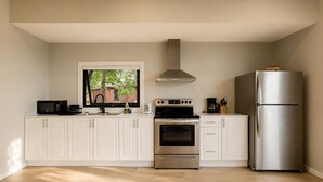 A modern kitchen with white cabinets, a stainless steel oven, a refrigerator, and a window overlooking a backyard. - Deluxe 3-Bedroom Cottage #12 (Nobel)