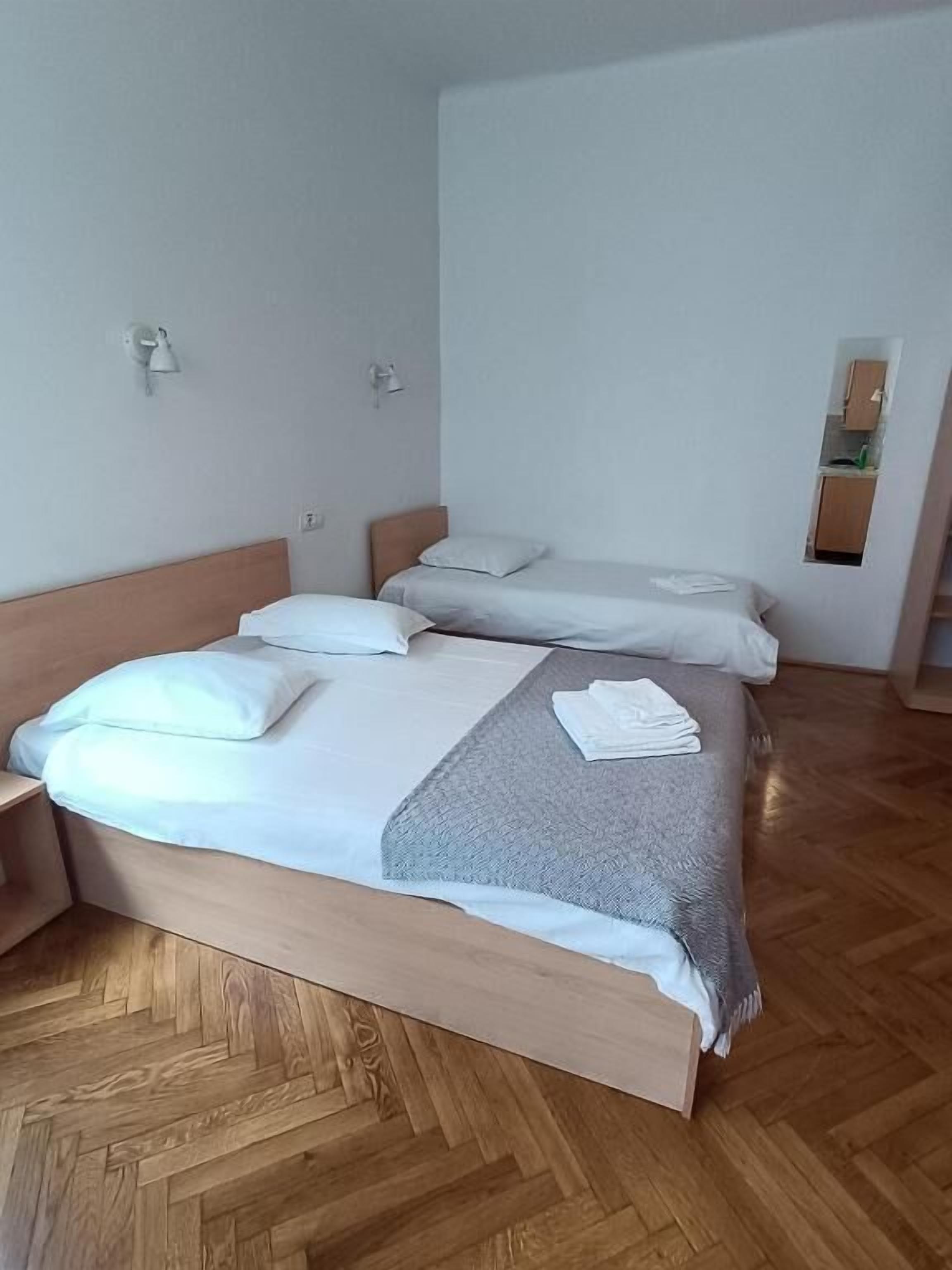 A hotel room with a large bed, a smaller bed, a wooden herringbone floor, and a small kitchenette visible through an open door.