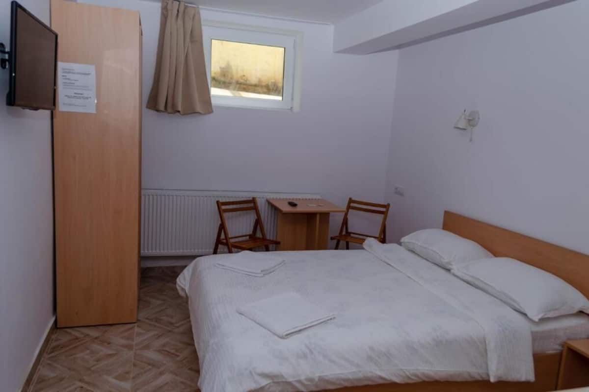 A bedroom with a bed, a desk, two chairs, a window with curtains, and a radiator.