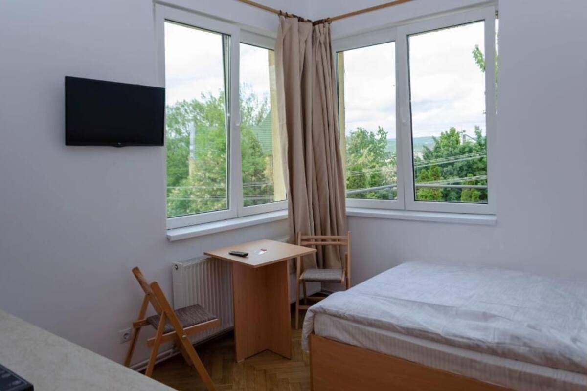 A bedroom with a bed, a desk, a chair, a TV, and large windows with curtains.