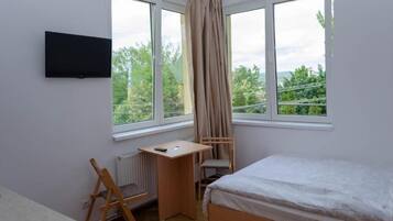 A bedroom with a bed, a desk, a chair, a TV, and large windows with curtains.