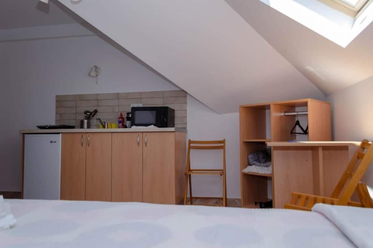 A compact room with a kitchenette, a bed, a chair, and a wooden wardrobe.