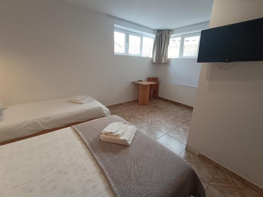 Ein Zimmer mit zwei Betten, einem kleinen Holzstuhl, einem an der Wand montierten Fernseher und einem Fenster mit Vorhängen.