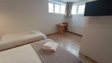 Ein Zimmer mit zwei Betten, einem kleinen Holzstuhl, einem an der Wand montierten Fernseher und einem Fenster mit Vorhängen.