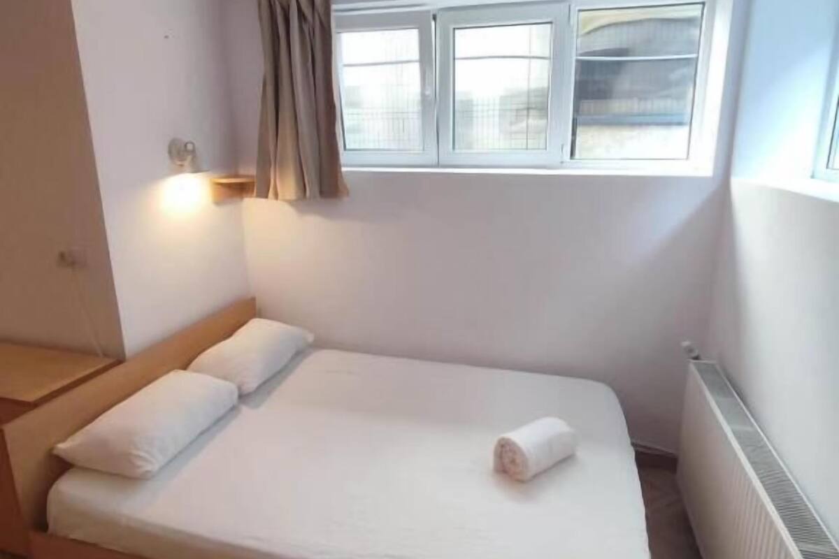 A single bed with a wooden frame and white bedding, a window with curtains, a wall-mounted light, and a radiator.