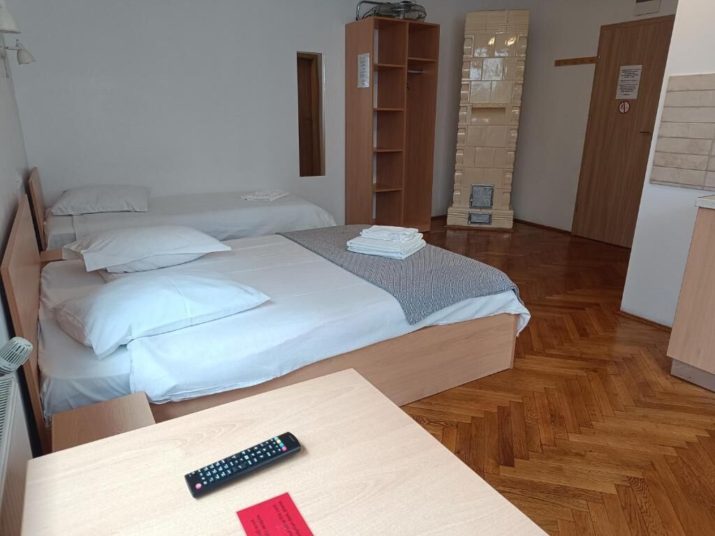 A bedroom with a bed, a wooden wardrobe, a small table with a remote control, and a wall-mounted shelf.