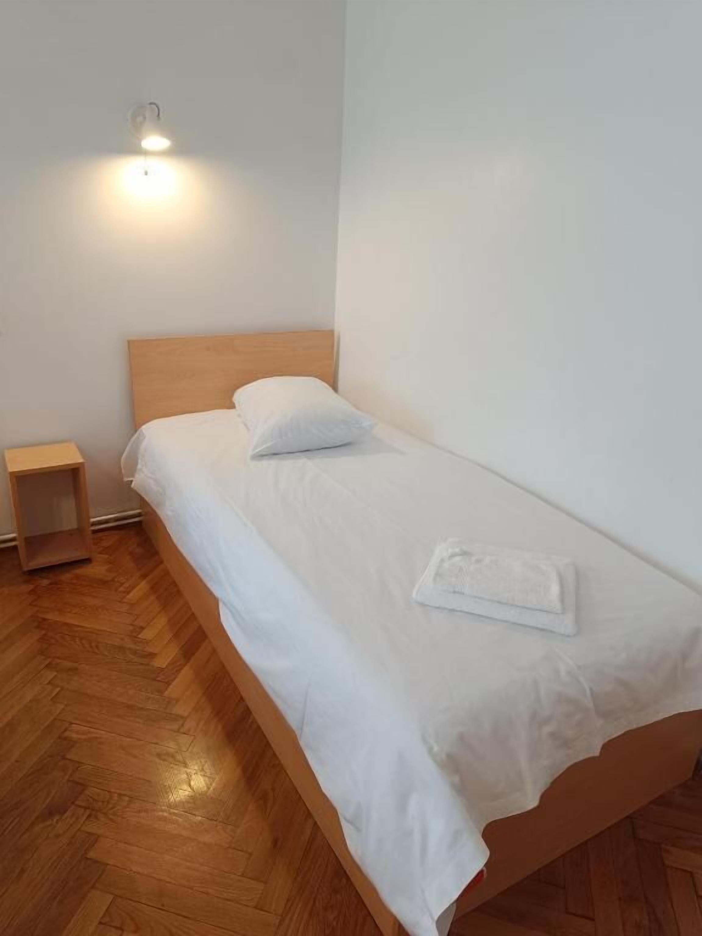 Una cama individual con cabecera de madera, una mesita de noche pequeña y una lámpara fijada a la pared.