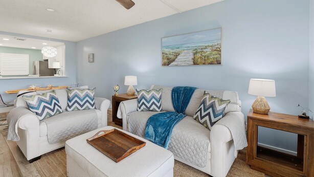 A living room with a ceiling fan, a painting of a beach scene, and a wooden side table with a lamp. - Sea Coast Condominium 517 (New Smyrna Beach)