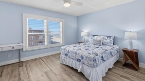 A bedroom with a bed, window, ceiling fan, bedside lamps, and a nightstand. - Sea Coast Condominium 517 (New Smyrna Beach)