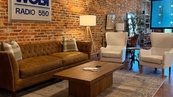 A living room with a brown leather sofa, two white armchairs, a wooden coffee table, and a brick wall.