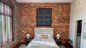 A bedroom with a brick wall, a large bed, two bedside tables, and a window with blinds. - Historic Media Loft in Downtown Columbus - Newly Renovated - Walk Everywhere (Columbus)