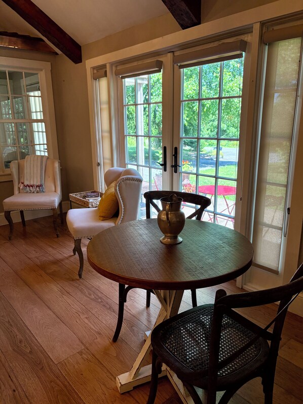 A room with a round table, two chairs, a bench, and a view through large glass doors to an outdoor area with a red table and chairs.