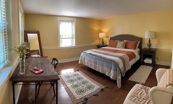 A bedroom with a bed, bedside tables, a nightstand, a chair, a table with a vase, and a window with blinds.