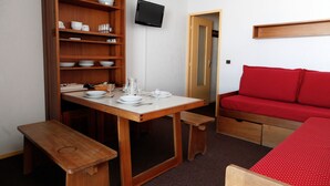 Quarto compacto com uma mesa de jantar de madeira posta para quatro pessoas, um sofá vermelho e uma televisão na parede.