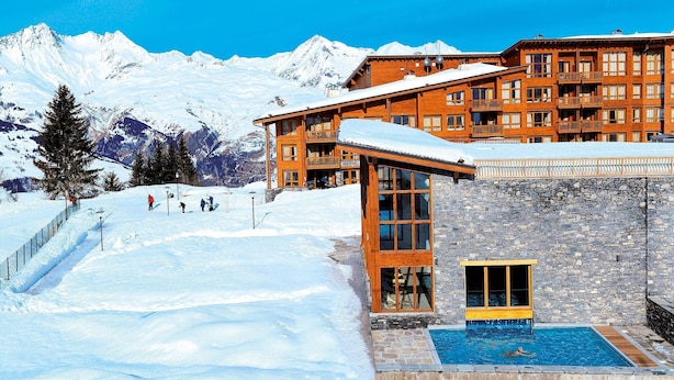 A hotel with a pool, ski slope, and snow-covered mountains in the background.