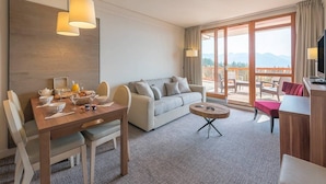 A hotel room with a dining area, a sofa, a TV, and a balcony with a view of the mountains.