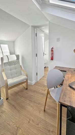 Ein moderner, gut beleuchteter Raum mit Holzboden, einem Esstisch mit Stühlen, einem an der Wand befestigten Feuerlöscher und einer Tür zu einem weiteren Raum.