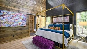 A bedroom with a four-poster bed, wooden walls, a large abstract painting, and a purple ottoman. - Luxury Cabin for Couples + Hot Tub + Fire Pit + Pets (Broken Bow)