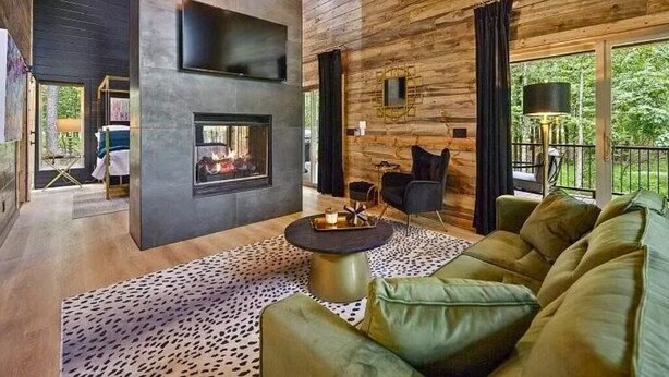 A modern living room with a fireplace, leather sofa, and a large window with a view of greenery. - Luxury Cabin for Couples + Hot Tub + Fire Pit + Pets (Broken Bow)