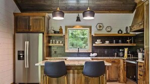 A kitchen with wooden cabinets, a refrigerator, a window, and two bar stools. - Luxury Cabin for Couples + Hot Tub + Fire Pit + Pets (Broken Bow)