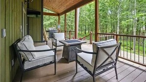 A covered outdoor area with wooden flooring, two cushioned chairs, a table, and a grill. - Luxury Cabin for Couples + Hot Tub + Fire Pit + Pets (Broken Bow)