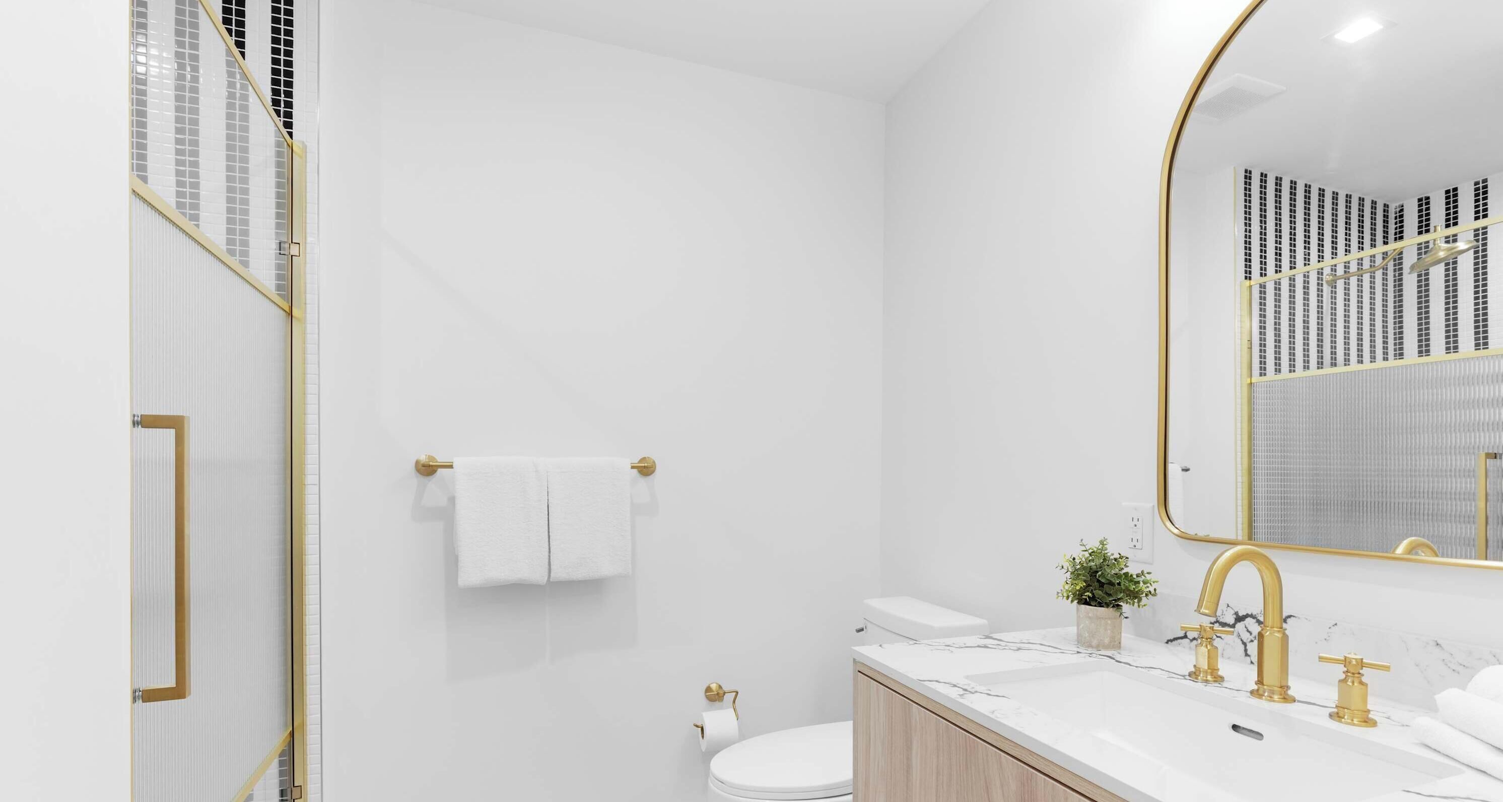 Une salle de bain moderne avec un lavabo blanc, des robinets dorés, un comptoir en marbre, des toilettes et un grand miroir avec un cadre doré.