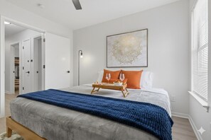 A bedroom with a bed, a bedside table with a tray, a wall-mounted light, and a framed artwork.