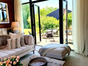 A modern living room with a sofa, a coffee table, and a view of an outdoor patio. - Lapworth Barn - Luxury Cottages (Lapworth)