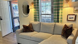 Ein Wohnzimmer mit einem Ecksofa, einem Teppich mit Muster, einem Fenster mit Vorhängen, einer Wandleuchte und einer Tür, die nach draußen führt.