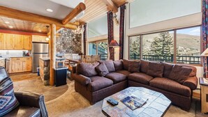 A cozy living room with a sectional sofa, a stone fireplace, and large windows offering a view of trees. - Lionshead Living | Steps from Gondola + Pool (Vail)