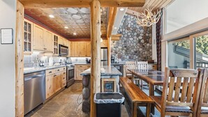 A kitchen with wooden cabinets, a stone wall, and a dining area with a bench and chairs. - Lionshead Living | Steps from Gondola + Pool (Vail)
