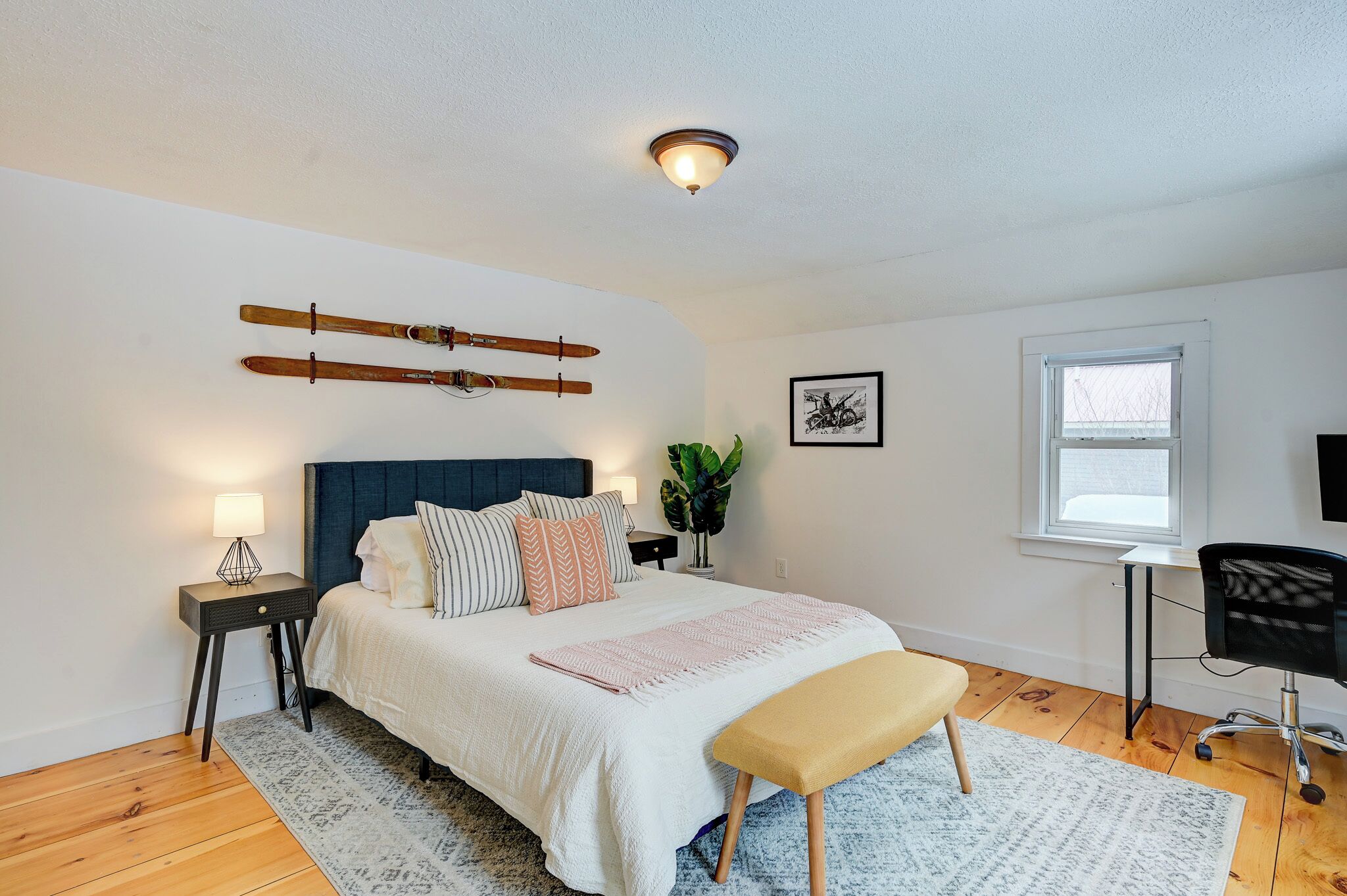 A bedroom with a bed, bedside tables, a desk, a chair, a window, and a ceiling light fixture.