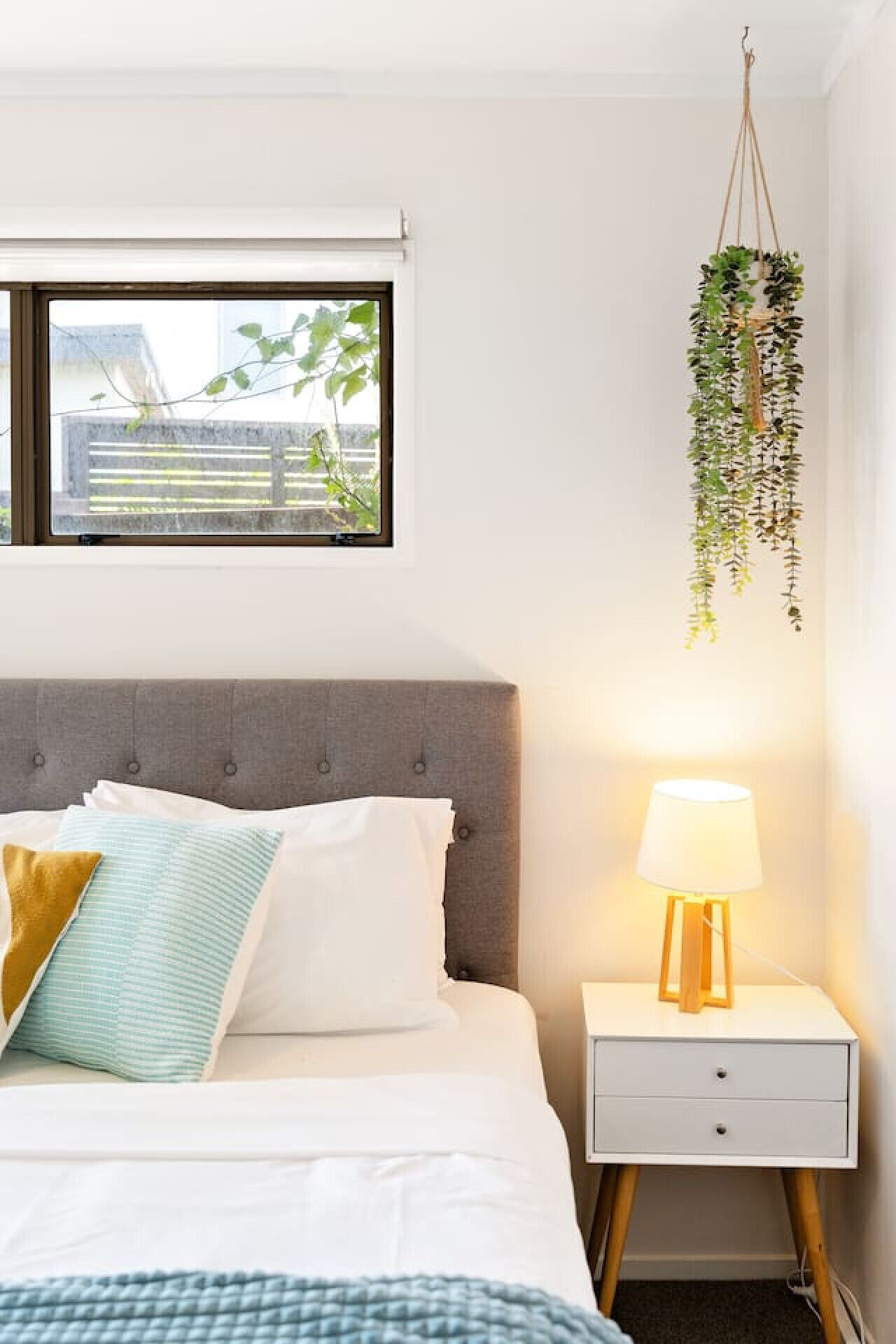 Une chambre à coucher avec un lit, une table de chevet, des plantes suspendues et une fenêtre.