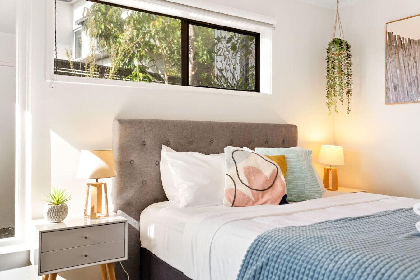 A bedroom with a grey upholstered headboard, a bed with white and patterned pillows, bedside lamps, a small plant on the nightstand, and a hanging plant.