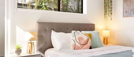 A bedroom with a grey upholstered headboard, a bed with white and patterned pillows, bedside lamps, a small plant on the nightstand, and a hanging plant.