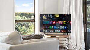 A television mounted on a stand in a room with a large window, a white couch, and a curtain. - Charming single-level home - just moments from the beach,  sun-soaked garden. (Auckland)
